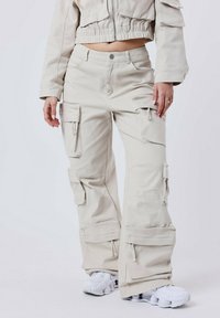 Pantalones cargo beige con múltiples bolsillos laterales, un ajuste relajado y costuras visibles. Combinados con calzado blanco.