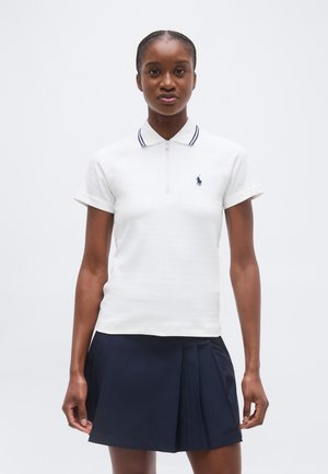 Młoda kobieta stoi, ubrana w białą koszulkę polo z krótkim rękawem i kołnierzykiem oraz granatową plisowaną spódnicę na jednolitym tle.