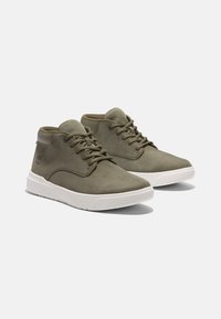 Botas chukka em pele verde-azeitona com solas em borracha branca, apresentando atacadores, uma língua em malha e um logótipo em relevo discreto na lateral.