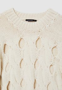 Maglione a maglia color crema con un motivo a trecce voluminose e sezioni traforate, colletto a costine e texture morbida. Etichetta del brand visibile.