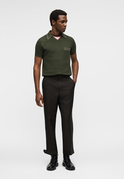 Άνδρας στέκεται με τα χέρια στις τσέπες φορώντας σκούρο πράσινο υφασμάτινο κοντομάνικο πουκάμισο, μαύρο παντελόνι και μαύρα μποτάκια με κορδόνια σε απλό φόντο.