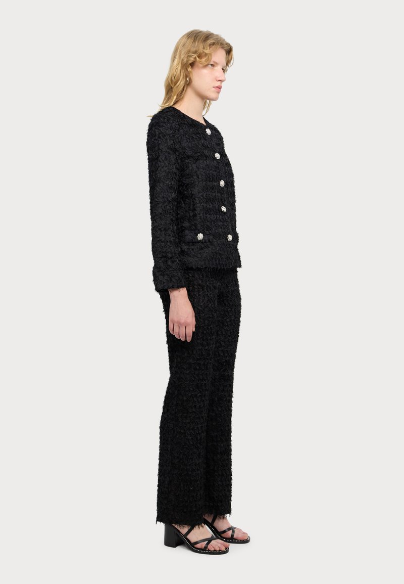 Zwarte geweven jas met textuur en zilveren knopen, gecombineerd met bijpassende textuurbroek. Model staat zijwaarts en toont de pasvorm van de outfit.