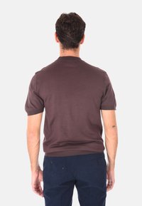 Uomo con capelli castani corti che indossa una camicia marrone a maniche corte e pantaloni blu scuro, in piedi rivolto di spalle su uno sfondo bianco.