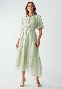 Vestido de capas en verde claro, hecho de una tela aireada. Presenta mangas cortas abullonadas, un cierre frontal con botones, un cinturón de tela con una hebilla circular y un dobladillo fluido.