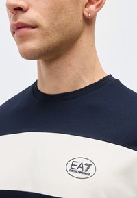 Κοντινό πλάνο ενός ατόμου που φοράει μπλε ναυτικό και άσπρο πουκάμισο EA7 Emporio Armani με ασημί κρίκο σκουλαρίκι στο αριστερό αυτί.