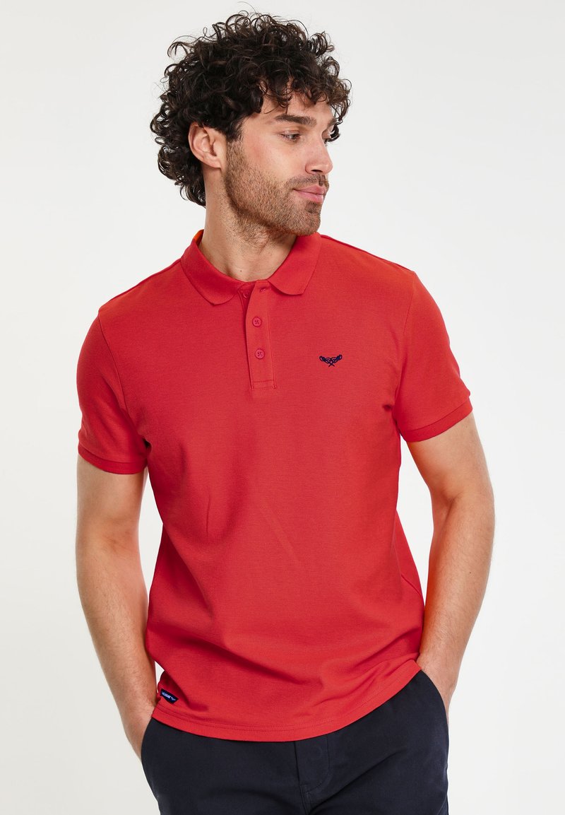 Camisa polo roja de algodón con cuello, tapeta de tres botones y detalle del logo en azul marino en el pecho. Mangas cortas, textura suave y diseño ajustado.