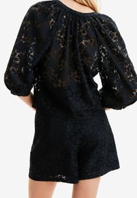 Top de encaje negro con mangas abullonadas y escote fruncido, combinado con pantalones cortos texturizados a juego, que presentan patrones florales y un efecto semitransparente.