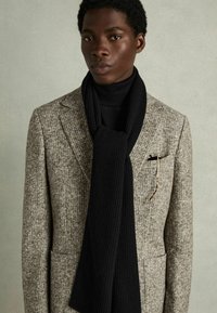 Grå og brun houndstooth-mønstrede blazer med spids klapper, parret med et ribbet sort tørklæde. En lommefyr og briller fuldender looket.