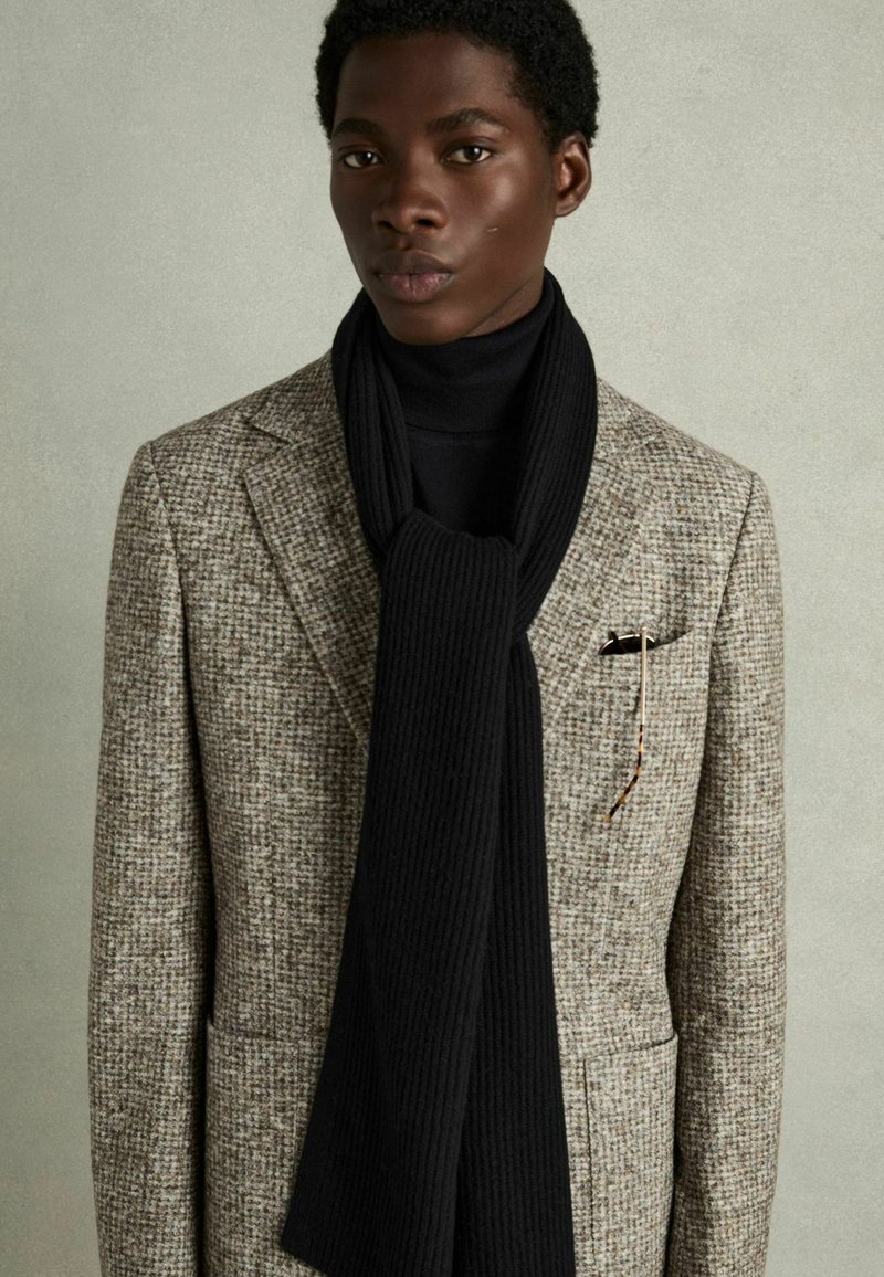Grå og brun houndstooth-mønstrede blazer med spids klapper, parret med et ribbet sort tørklæde. En lommefyr og briller fuldender looket.