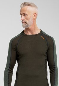 Langærmet thermal top i mørkegrøn og sort, med strukturerede grønne skuldre, tætsiddende pasform og subtilt brandlogo på brystet.