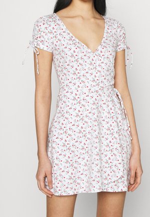 Woman wearing a white short-sleeve wrap dress with red and green small floral print and adjustable sleeve ties.