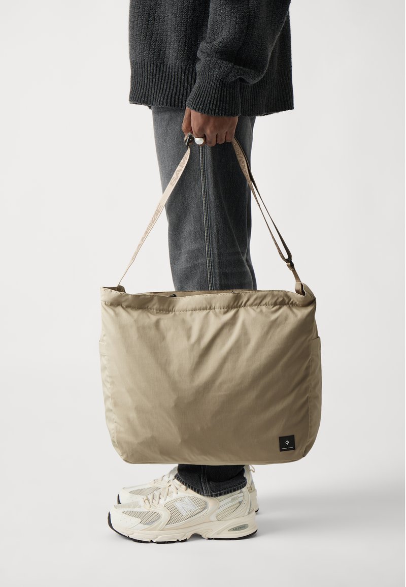Beige isolert bag med rektangulær form, bæresnor og logo-patch; båret av en person i mørk jeans og hvite joggesko.