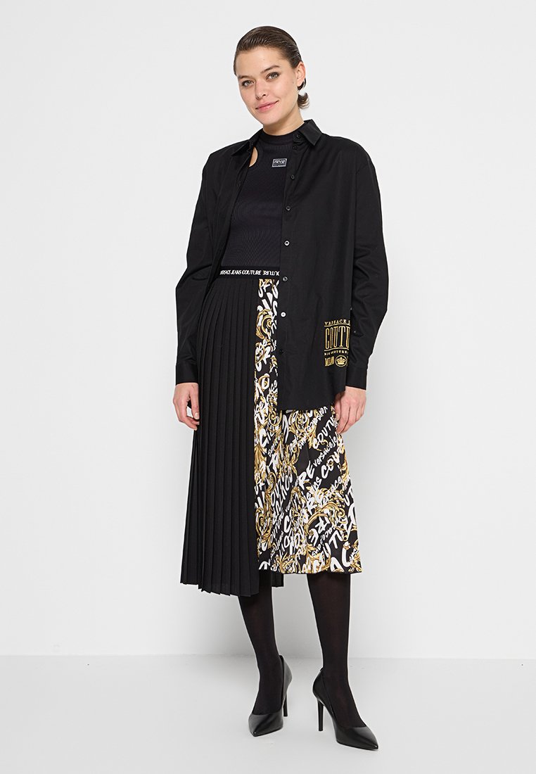 Schwarzes Hemd mit Knöpfen und Logo, kombiniert mit einem schwarzen Faltenrock und einem bedruckten Rock mit goldenen und weißen Mustern. Schwarze Absatzschuhe.