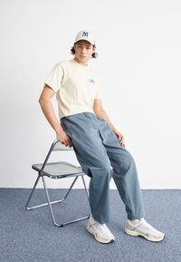 Beige bomull t-shirt med blå grafik, grå avslappnade byxor och vita sneakers, sittande på en kromad hopfällbar stol. Bakgrunden är enfärgad.