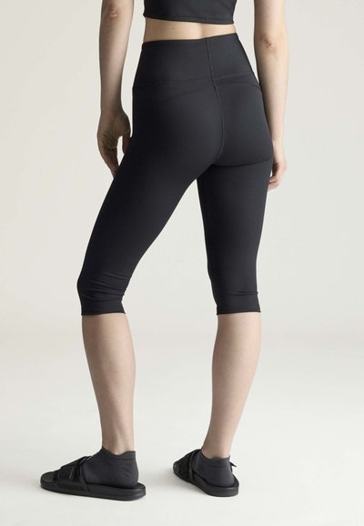 Person iført sorte korte leggings, grå sokker og sorte slid-sandaler, stående mod en ensartet lys baggrund, set bagfra.