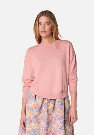 Lichtroze sweater met een golvende rand en een losse pasvorm. Beschikt over een zachte textuur en lange mouwen, gecombineerd met een rok met bloemenpatroon.