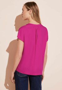 Blusa fucsia a maniche corte con dettaglio plissettato sul retro. Materiale leggero con una texture liscia. Abbinata a jeans in denim blu scuro.