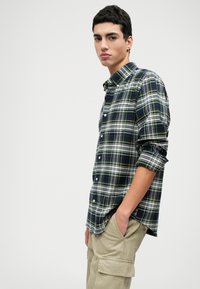 Rutet knappeskjorte i marineblå, grønn og hvit; lange ermer opprullet; laget av lett stoff; har klassisk krage og frontknapper.