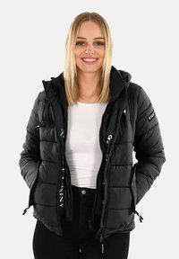 Šypsena moteris su šviesiais plaukais, vilkinti juodą pūkinę striukę virš baltos palaidinės ir juodų kelnių, rankos kišenėse.