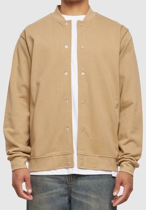 Beige Bomberjacke mit Knopfleiste, getragen über einem weißen Hemd und blauen Jeans, gezeigt vom Hals bis zur Taille, Arme entspannt seitlich hängend.