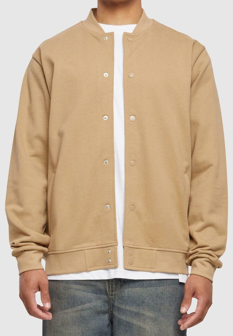 Veste bomber beige boutonnée portée sur une chemise blanche avec un jean bleu, visible du cou à la taille, les bras détendus le long du corps.