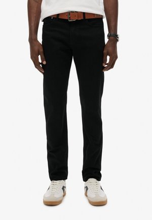 Mand iført sorte jeans, hvide sneakers med sorte striber, et brunt bælte og et sort armbåndsur mod en hvid baggrund.