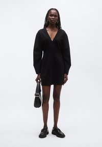 Μαύρο μίνι φόρεμα με V-λαιμόκοψη, μακριά φαρδιά μανίκια και λεία υφή. Συνδυασμένο με μαύρα loafers και δομημένη τσάντα χειρός.