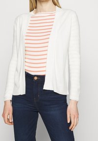 Cárdigan blanco con abertura frontal, tejido texturizado, mangas largas sobre una camiseta de rayas en melocotón y blanco, combinado con vaqueros azul oscuro.