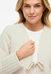 Giacca trapuntata beige chiaro con chiusura a nastro, caratterizzata da una vestibilità comoda e un tessuto morbido e testurizzato. Indossata sopra un top bianco a coste.