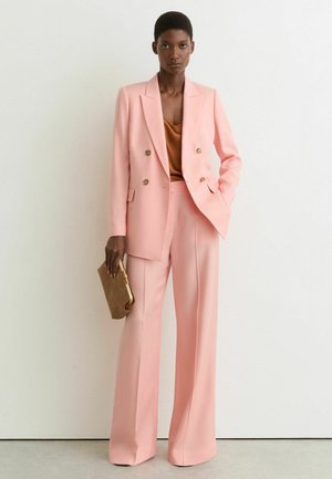 Žena nosící růžové dvouřadé sako s širokými kalhotami a hnědou blůzou, držící hnědou psaníčko, stojící před jednobarevnou bílou stěnou.