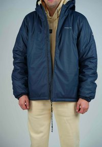 Doudoune marine avec capuche, poignets élastiques et fermeture éclair à l'avant. Portée sur un sweatshirt beige, associée à un pantalon de couleur crème.