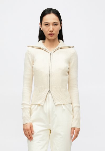 Pull crème zippé avec col haut, poignets et ourlet côtelés, confectionné en maille douce, présentant une coupe ajustée et une texture subtile.