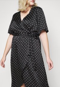 Schwarzes Wickelkleid aus Satin mit weißen Punkten, kurzen Ärmeln und einem Bindegürtel in der Taille, mit einem tiefen V-Ausschnitt und asymmetrischem Saum.