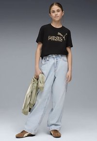 Svart t-shirt med leopardmönster, löst sittande ljusblå jeans och bruna sneakers. Håller en ljusgrön jacka. Enfärgad grå bakgrund.