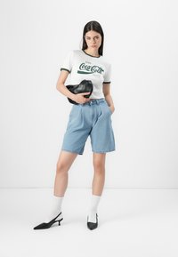 White t-shirt with green accents featuring a Coca-Cola graphic, paired with light blue denim shorts, black high heels, and white socks.