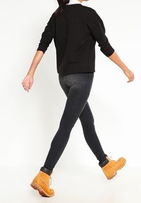 Jersey negro de gran tamaño, vaqueros oscuros de cintura alta y botas amarillas con un diseño de cordones. Modelo caminando, mostrando el ajuste y contraste del atuendo.