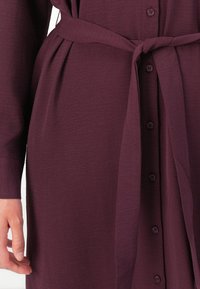 Maroon dress with a button-down front, long sleeves, and a waist tie. The texture is slightly crinkled, adding visual interest.