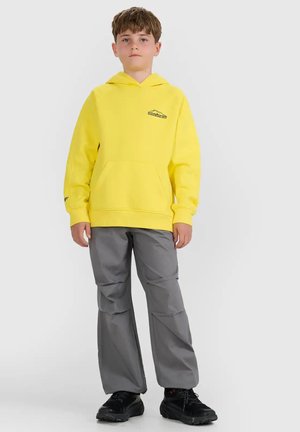 Niño con una sudadera con capucha amarilla brillante, pantalones grises y zapatillas negras, de pie frente a un fondo blanco liso.