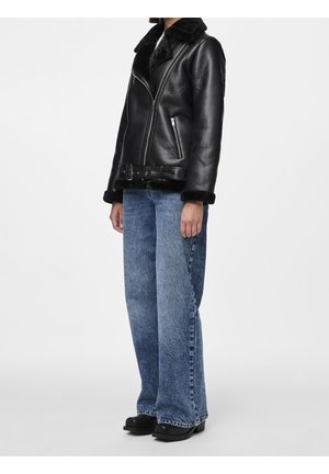 Osoba ubrana w czarną kurtkę ze sztucznego skóropodobnego kożucha z suwakiem i paskiem, do tego luźne, niebieskie jeansy z efektem sprania oraz czarne buty.