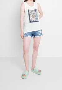 Biała bluzka bez rękawów z centralnym niebiesko-pomarańczowym geometrycznym wzorem, połączona z przetartymi szortami dżinsowymi i miętowymi sandałami na platformie.