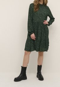 Vestido verde con estampado floral, mangas largas, cintura fruncida y falda de volantes. Combinado con botines negros con suelas gruesas y laterales elásticos.
