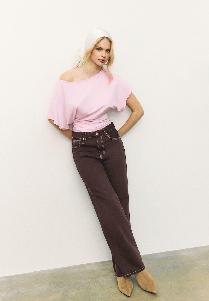 Jeune femme appuyée contre un mur, portant un haut rose à épaules dénudées, un pantalon marron foncé, des bottes pointues couleur beige et un foulard blanc.