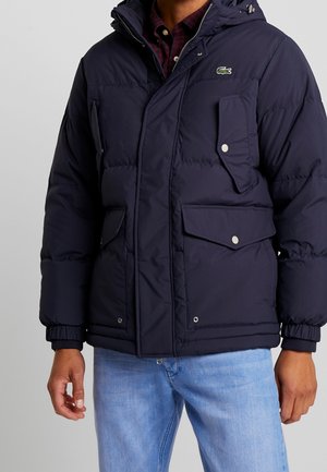 Man in een marineblauwe gewatteerde jas met klepzakken en het Lacoste-logo, gecombineerd met lichtblauwe spijkerbroek en een rood geruit overhemd eronder.