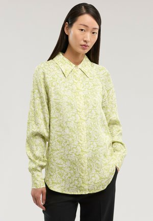 Femme aux longs cheveux noirs portant un chemisier à manches longues vert clair à motifs avec un large col et un pantalon noir, debout devant un fond uni.