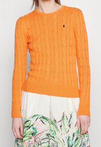 Nainen, jolla on yllään kirkkaan oranssi joustinneulepusero ja valkoinen hame, jossa on värikkäitä abstrakteja vihreitä ja vaaleanpunaisia kuvioita.