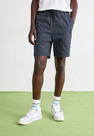 Mężczyzna w czarnych szortach z troczkami, białych skarpetkach z dwiema paskami oraz białych trampkach stojący na zielonej podłodze przy białej ścianie.