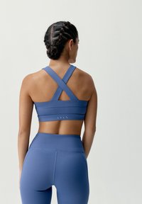 Modrá športová podprsenka s prekriženým dizajnom na chrbte, vyrobená z elastického materiálu. K outfitu sa hodia vysoké modré legíny.