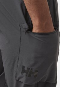 Szare, lekkie szorty z tkaniny z zamykana boczną kieszenią i błyszczącym czarnym logo na nodze. Gładka tekstura, sportowy fason.