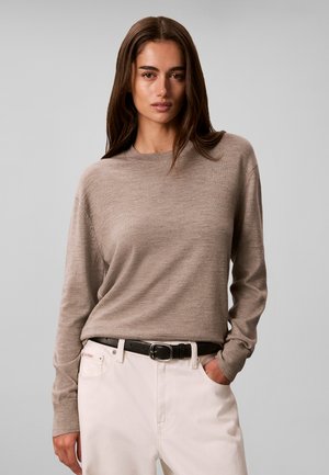 Kvinde med langt brunt hår iført en beige sweater stoppet ind i hvide bukser med sort bælte, stående mod en ensfarvet grå baggrund.
