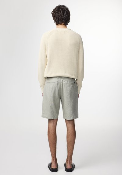 Helles beiges Strickpullover mit langen Ärmeln, kombiniert mit hellgrauen Shorts. Die Shorts haben eine lockere Passform und eine dezente Textur. Schlichte schwarze Sandalen.